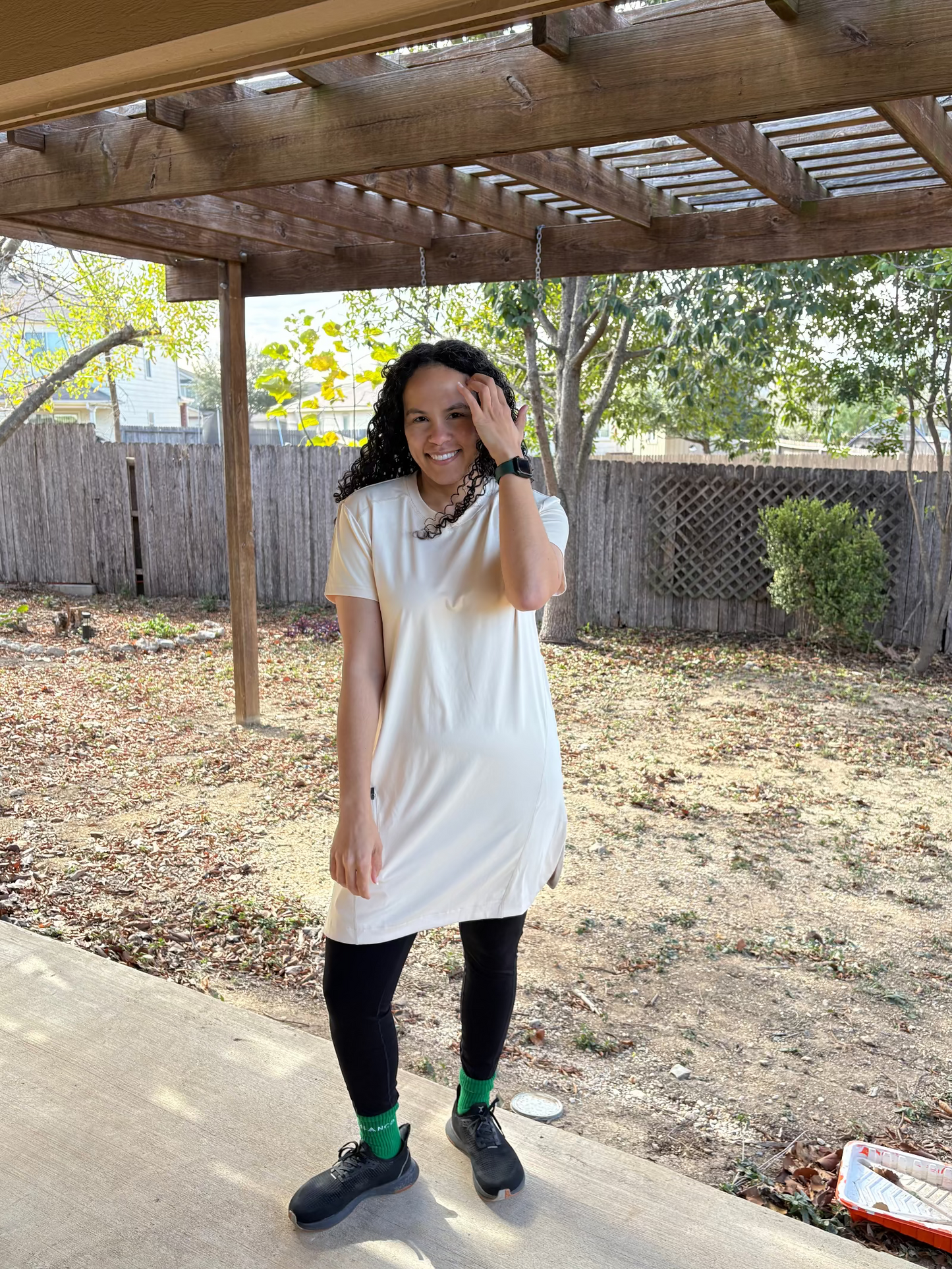 Woman in a white dress standing under a wooden pergola with trees and a fence in the background.