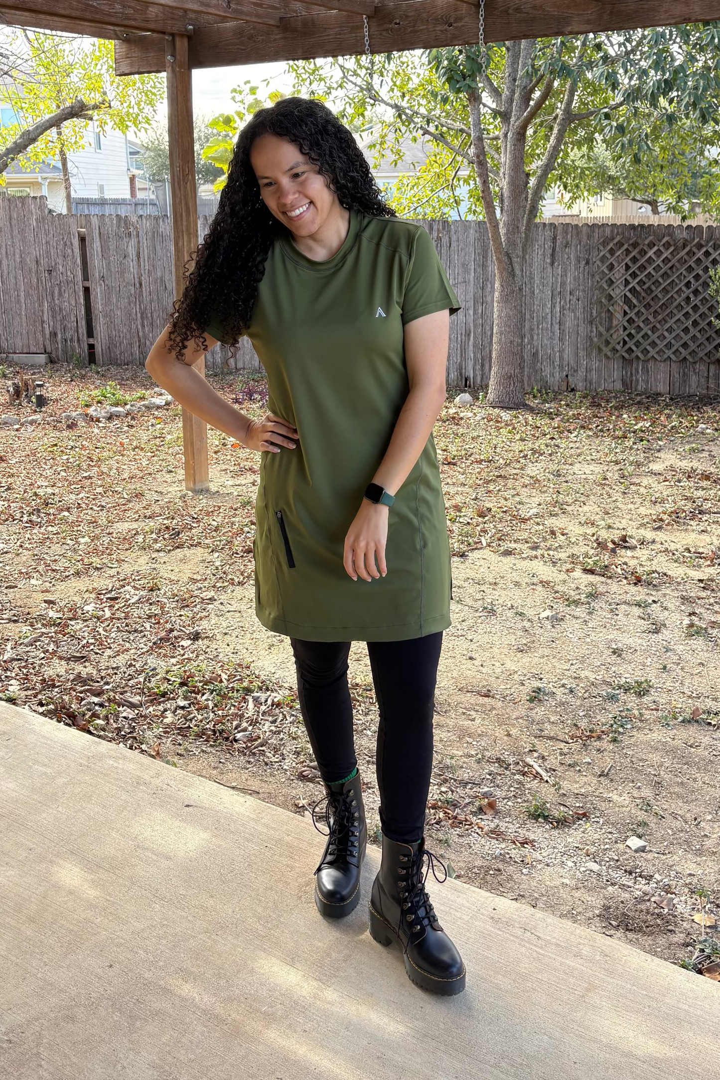 Woman in a green dress standing under a wooden pergola with trees and a fence in the background.