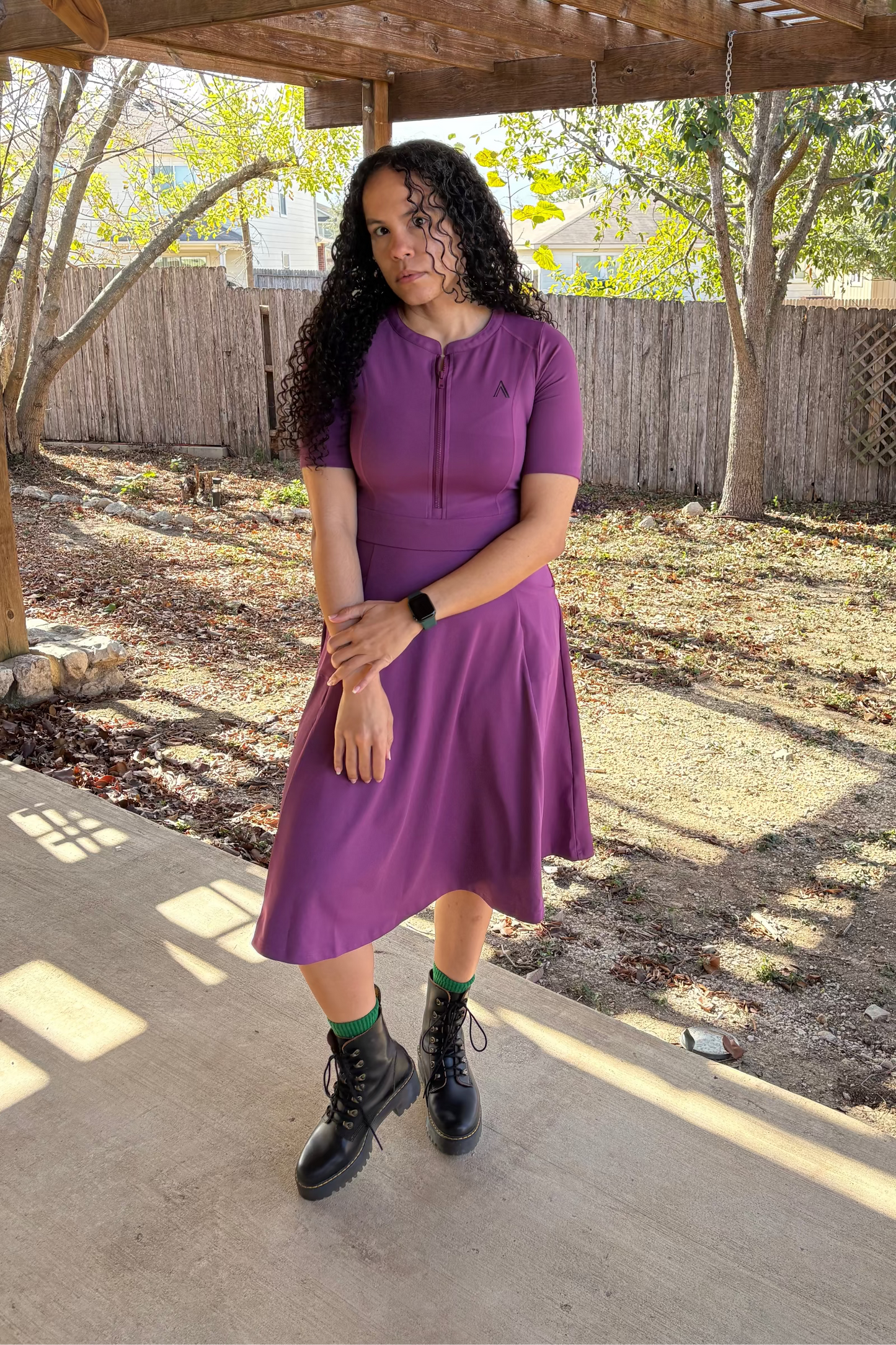 Woman in a purple dress standing under a wooden pergola with trees and a fence in the background.