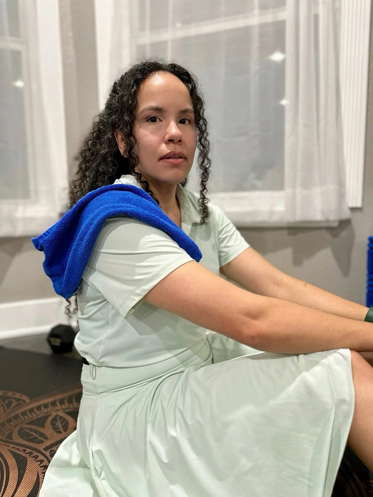 Woman sitting on a yoga mat with a blue towel draped over her shoulder, in front of a window.